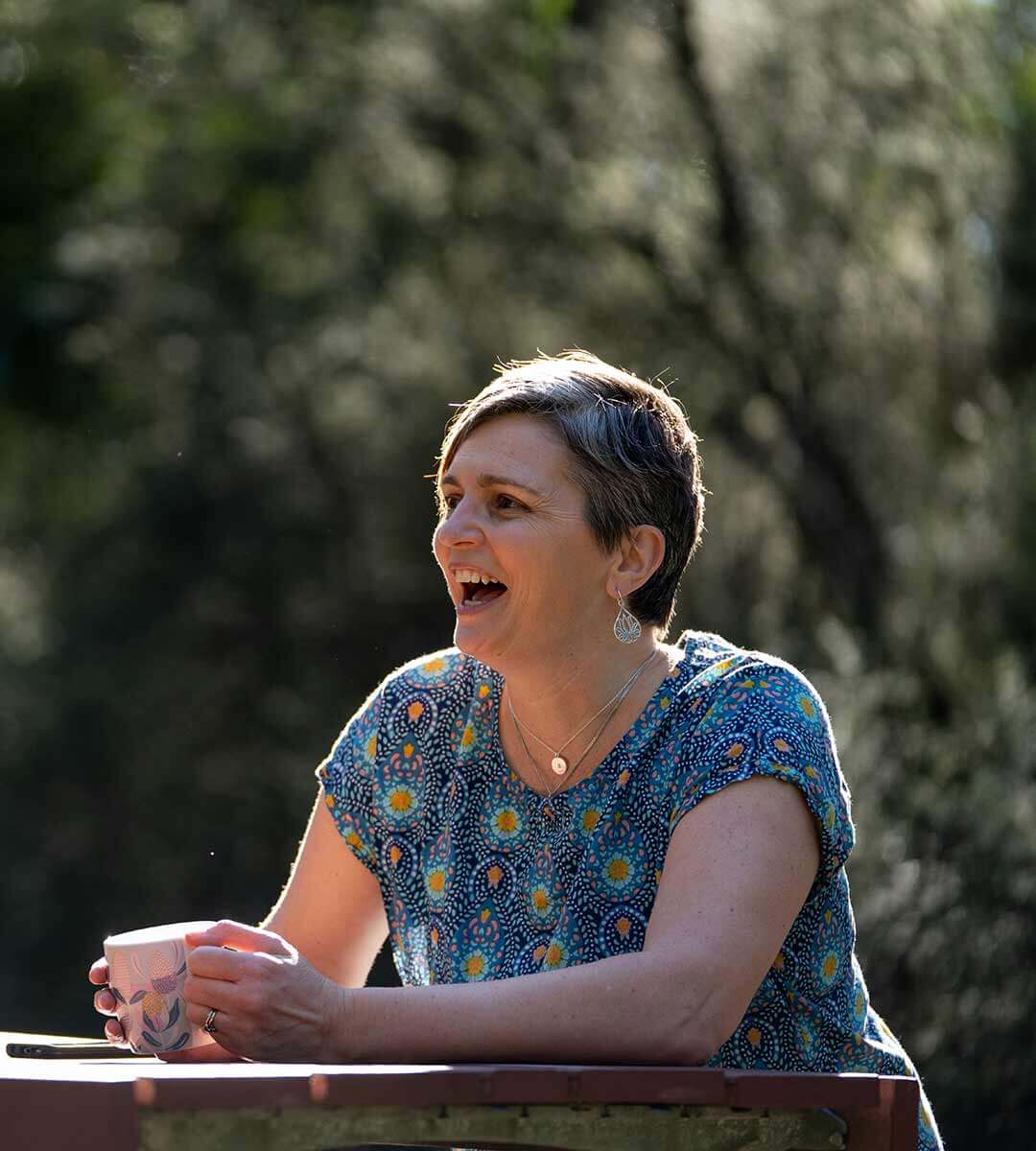 Mandy Mercuri, owner and mindfulness coach, laughing at a bench outside in the sun.