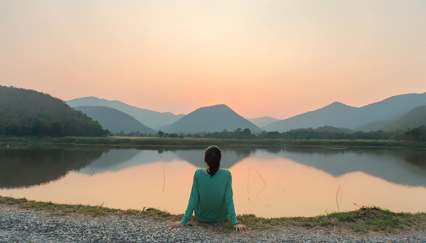 Woman sitting by a lake with her back to the camera, facing the mountains in mindfulness.