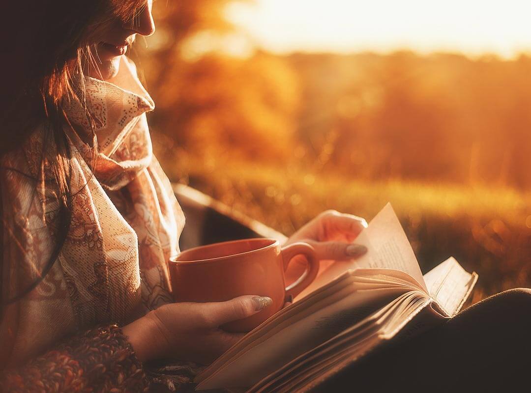 A woman sitting in the autumn sun, wrapped up warm with a book on her lap.